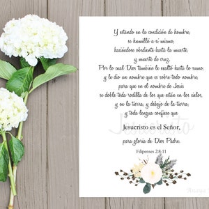 May include: A white floral print with text in Spanish, featuring a verse from Philippians 2:8-11, is displayed on a wooden surface. Two large white hydrangea blooms with green leaves are placed on the left side of the image.