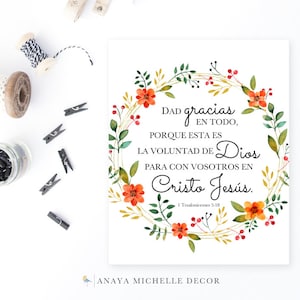 May include: A square print with a floral wreath design. The wreath features orange flowers, green leaves, and gold accents. The print contains Spanish text, including "Dad gracias en todo, porque esta es la voluntad de Dios para con vosotros en Cristo Jesús."