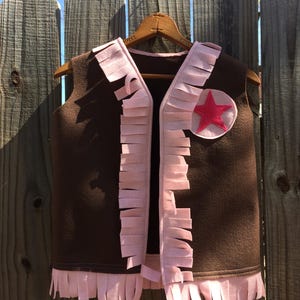 May include: A brown felt vest with pink fringe trim and a pink felt star on the front. The vest is hanging on a wooden hanger.