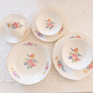 May include: A collection of vintage white porcelain dishware with gold trim. The set includes a teacup and saucer, bowls, and plates, all adorned with a floral pattern in pink, blue, and yellow. The edges of the plates and bowls have a scalloped design.