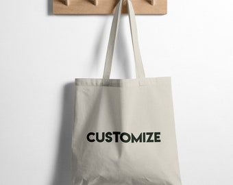 Könnte beinhalten: Eine schlichte weiße Tragetasche mit schwarzem Text "CUSTOMIZE" hängt an einem Holzgestell mit Haken.