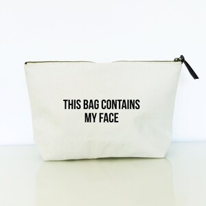 May include: A white canvas zippered pouch with black text that reads "This bag contains my face."