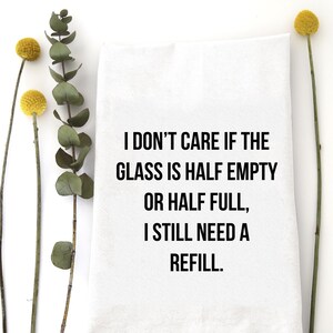 Puede incluir: Paño de cocina blanco con el texto "I DON'T CARE IF THE GLASS IS HALF EMPTY OR HALF FULL, I STILL NEED A REFILL." La toalla está rodeada de detalles florales amarillos y verdes.