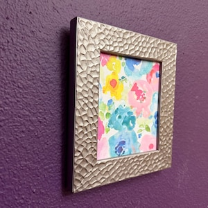 May include: A square picture frame with a metallic silver finish and a textured, hammered surface. The frame contains a watercolor painting featuring a vibrant floral pattern with overlapping petals in shades of yellow, pink, and blue.