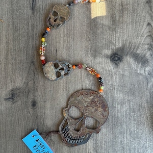 May include: A rusty metal skull garland with orange, red, and black beads. The garland is hanging on a wooden surface. The tag attached to the garland reads "WINDY CITY Bells".