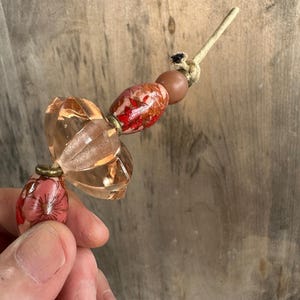 May include: A handmade beaded ornament featuring a combination of clear, faceted, and floral-patterned beads in shades of pink, red, and brown. The ornament is strung on a beige cord and knotted at the top.