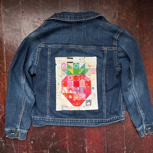May include: A blue denim jacket with a patchwork strawberry design on the back. The strawberry is made of colorful fabric squares.