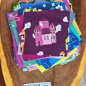 Puede incluir: Una pila de cuadrados de tela de colores con un cuadrado morado en la parte superior que presenta un castillo rosa y fantasmas. Los cuadrados de tela están dispuestos en una pila sobre una superficie de madera. El texto "POSTCARD QUILTS" es visible en una pegatina en la esquina inferior derecha de la imagen.