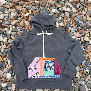 Puede incluir: Una sudadera con capucha gris con un gran bolsillo de patchwork en la parte delantera. El bolsillo está hecho de varias telas coloridas con un patrón floral y geométrico. El bolsillo está cosido a la sudadera con un hilo blanco contrastante.