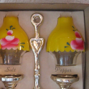 May include: A set of salt and pepper shakers in the shape of lampshades. The shakers are yellow with pink and green painted flowers. The shakers are in a box with the words "Salt" and "Pepper" printed on the bottom.