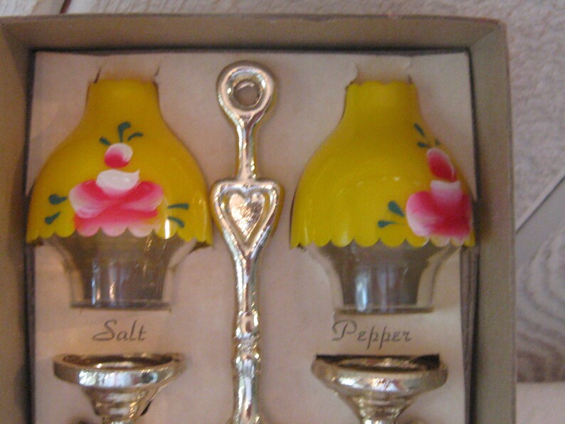 May include: A set of salt and pepper shakers in the shape of lampshades. The shakers are yellow with pink and green painted flowers. The shakers are in a box with the words "Salt" and "Pepper" printed on the bottom.