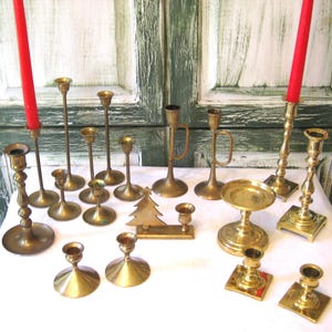 May include: A collection of brass candle holders in various shapes and sizes, including tall, slender designs and shorter, wider bases. Two red candles are lit, adding a pop of color. A brass Christmas tree is also present.