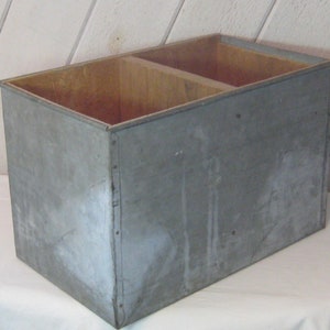 Puede incluir: Una caja de almacenamiento grande, rectangular, de metal con una tapa de madera. La caja está dividida en dos compartimentos. La caja está pintada de color gris claro y tiene un aspecto desgastado y vintage.