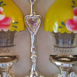 May include: A set of salt and pepper shakers with a yellow base and a floral design. The shakers are labeled "Salt" and "Pepper".