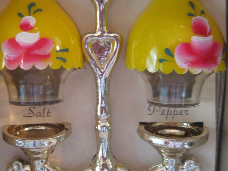 May include: A set of salt and pepper shakers with a yellow base and a floral design. The shakers are labeled "Salt" and "Pepper".