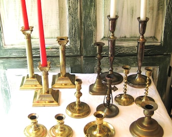 May include: A selection of antique brass and silver candlesticks in various styles. Some hold red candles, others white. The candlesticks are displayed on a white surface, set against a distressed green door.