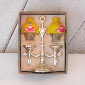 May include: A vintage salt and pepper shaker set in the shape of lamp posts with yellow glass shades and pink floral accents. The shakers are attached to a silver metal stand with a heart-shaped design. The box reads "Salt" and "Pepper".