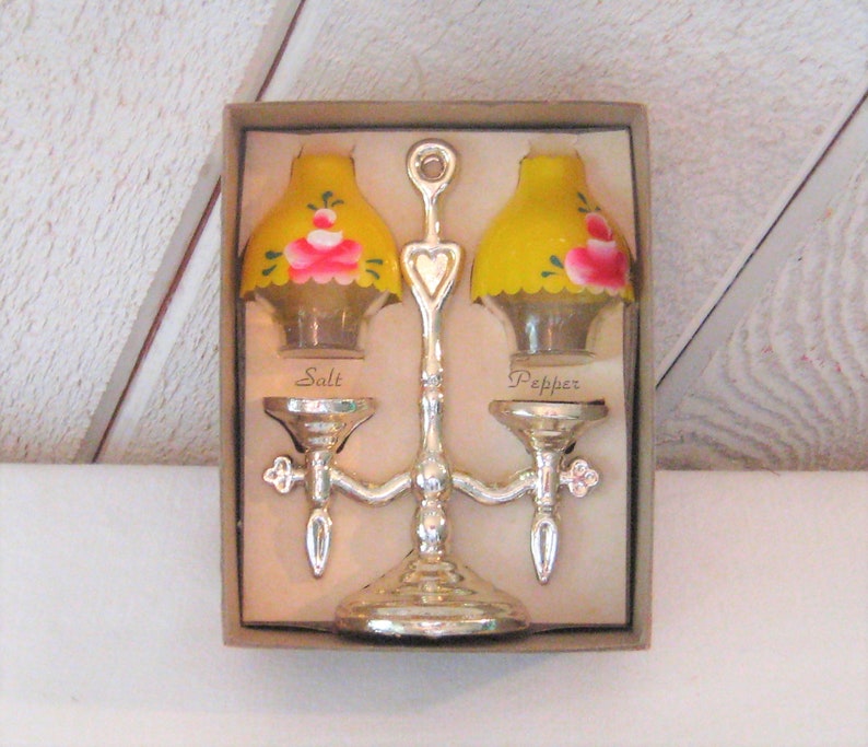 May include: A vintage salt and pepper shaker set in the shape of lamp posts with yellow glass shades and pink floral accents. The shakers are attached to a silver metal stand with a heart-shaped design. The box reads "Salt" and "Pepper".