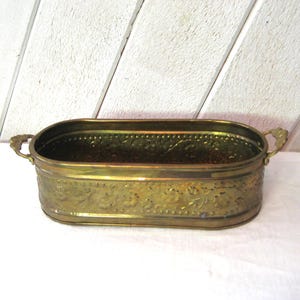 Pode incluir: Um vaso de latão vintage com formato oval e detalhes florais ornamentados. O vaso possui duas alças decorativas e provavelmente é usado para exibir plantas ou flores. O latão tem uma pátina quente e envelhecida. Medidas em centímetros.