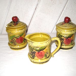 May include: A vintage ceramic kitchen set in a yellow-green hue, featuring two lidded jars and a creamer. Each piece is adorned with a fruit motif, including red apples and purple grapes, and a red apple knob on the jar lids.