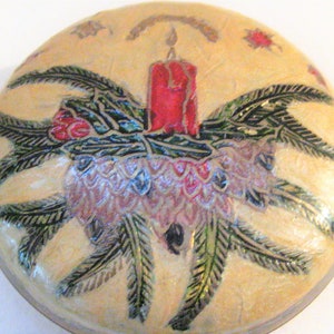 May include: Round, cream-colored decorative box with a Christmas theme. The lid features a red candle surrounded by green pine needles, red berries, and a pinecone. Gold accents and small star details complete the festive design.