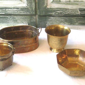 May include: Four brass planters of different shapes and sizes. The largest planter is oval and has a lip around the top. The second largest planter is round and has a flared lip. The smallest planter is round and has a flared lip and three feet. The fourth planter is octagonal and has a flared lip.