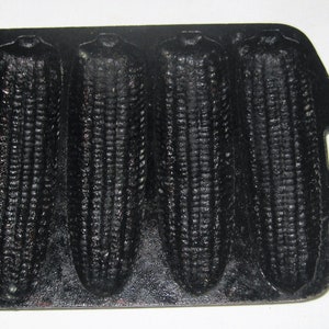 May include: A black cast iron cornbread pan with six individual molds shaped like ears of corn. The pan has a handle on the side.