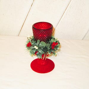 May include: A red glass candle holder with a textured surface, adorned with a festive holly and berry wreath. The candle holder has a round base and a decorative design, perfect for holiday decor.