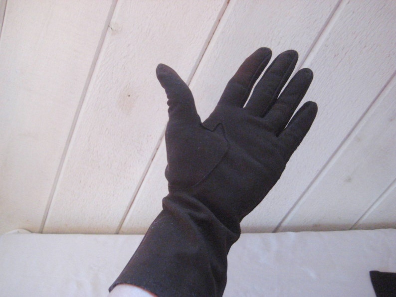 May include: A pair of long black gloves made of a soft fabric. The gloves have a seam down the back of the hand and a small patch on the back of the hand.