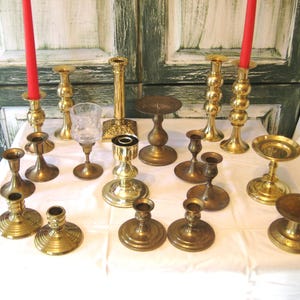 Puede incluir: Una colección de candelabros y portavelas de latón de varios tamaños y diseños. Dos velas rojas están encendidas en soportes altos. La colección incluye una copa de cristal y varios estilos de portavelas.