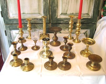 May include: A selection of brass candlesticks and candle holders in various styles. Two red candles are lit in tall holders. The collection includes a glass goblet and several different styles of candle holders.
