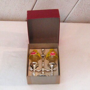 May include: A pair of yellow glass salt and pepper shakers with pink floral designs, mounted on a silver stand. The shakers are in a red and white box.