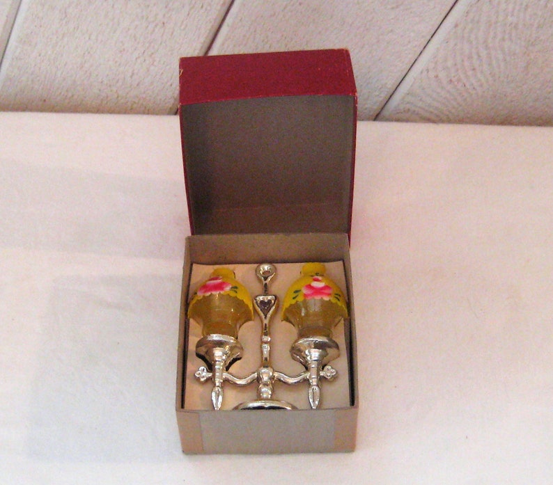 May include: A pair of yellow glass salt and pepper shakers with pink floral designs, mounted on a silver stand. The shakers are in a red and white box.
