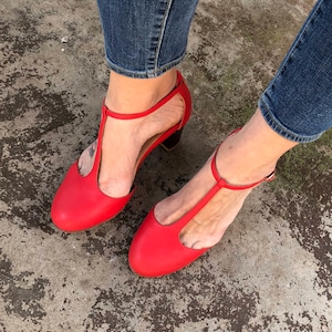 May include: A pair of red leather heeled shoes with ankle straps. The shoes have a t-strap design and are worn by a person wearing blue jeans.