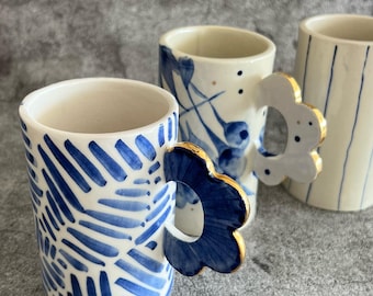以下が含まれることがあります： 青と白のデザインのセラミックマグ3つ。前面のマグは、金色のトリムと青い筆のストロークパターンが付いた青い花のハンドルが付いています。中央のマグは、金色のトリムと青い花のアクセントが付いた白い花のハンドルが付いています。