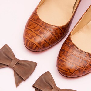 May include: Two pairs of brown leather bow ties. The bow ties are made of a textured material that resembles crocodile skin. The bow ties are on a pink background.