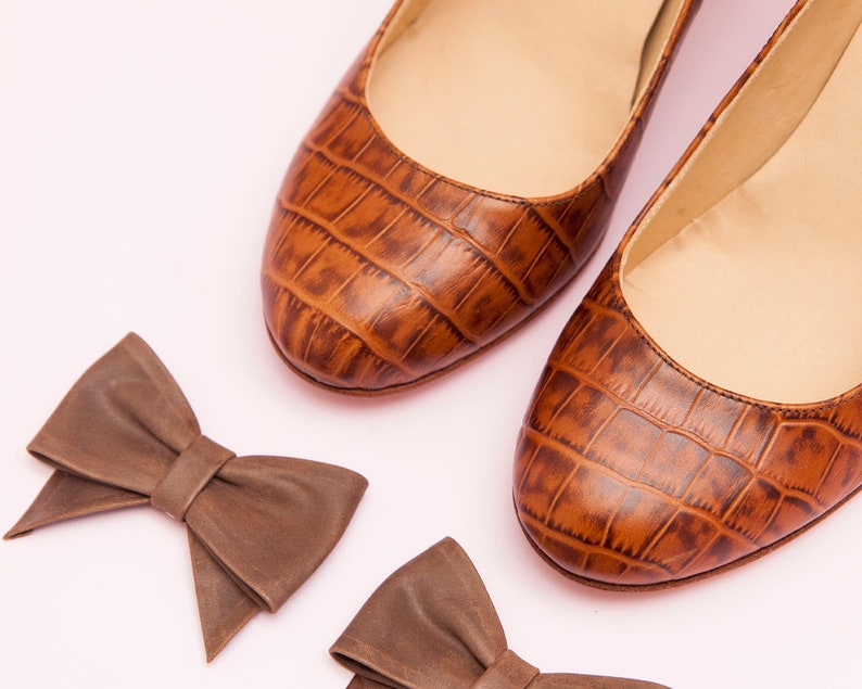 May include: Two pairs of brown leather bow ties. The bow ties are made of a textured material that resembles crocodile skin. The bow ties are on a pink background.