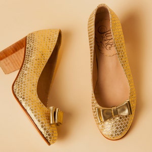 May include: A pair of gold shoes with a polka dot pattern and a bow on the toe. The shoes have a brown heel and the brand name "June" is printed on the inside of the shoe.
