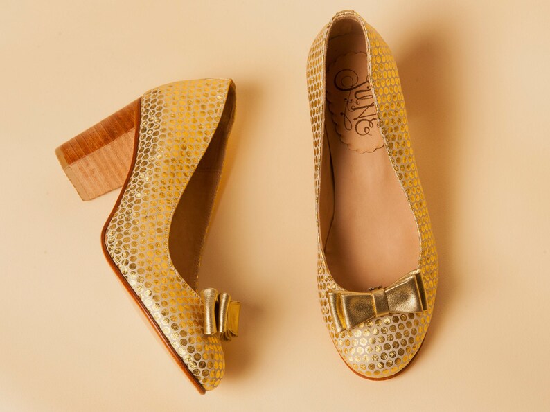 May include: A pair of gold shoes with a polka dot pattern and a bow on the toe. The shoes have a brown heel and the brand name "June" is printed on the inside of the shoe.