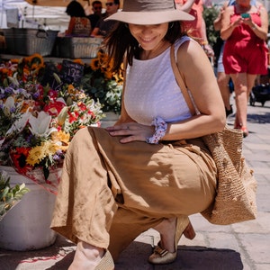 May include: A woman wearing a white sleeveless top, a brown hat, and tan wide-leg pants. She is sitting on a brick walkway with a straw bag and a bouquet of flowers in the background.