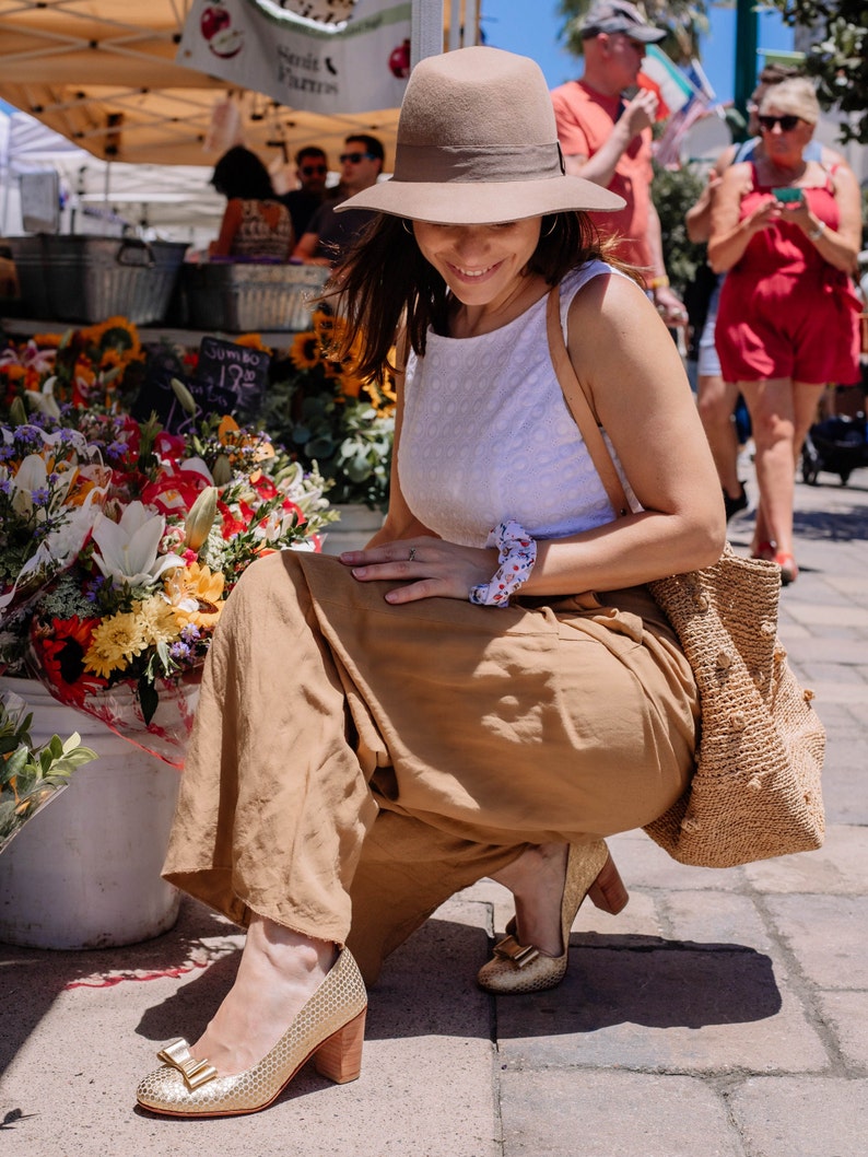 May include: A woman wearing a white sleeveless top, a brown hat, and tan wide-leg pants. She is sitting on a brick walkway with a straw bag and a bouquet of flowers in the background.