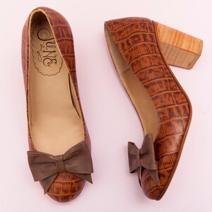 May include: A pair of brown leather heeled shoes with a crocodile print and a brown bow on the front. The shoes have a wooden heel.