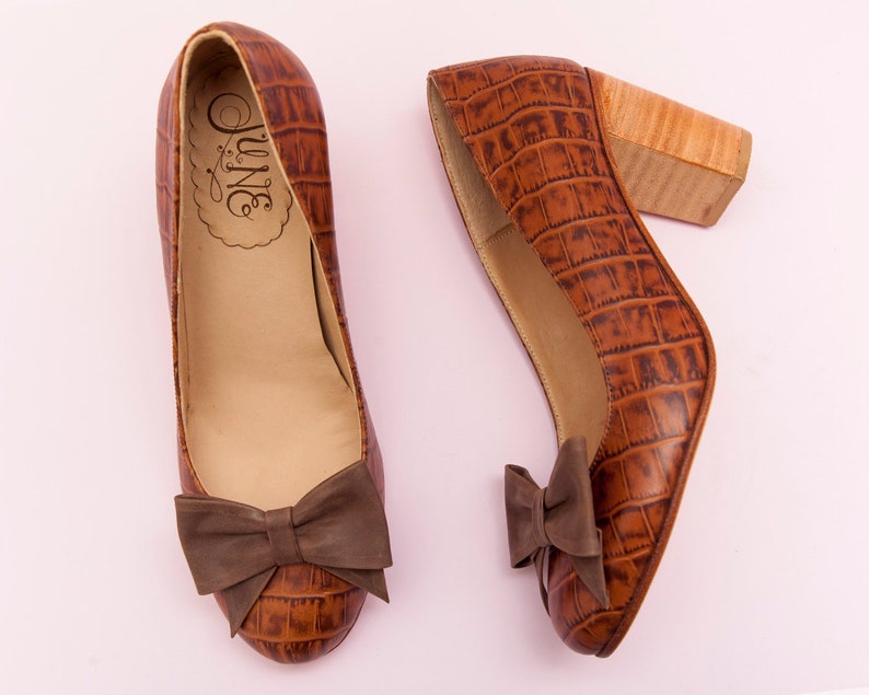 May include: A pair of brown leather heeled shoes with a crocodile print and a brown bow on the front. The shoes have a wooden heel.