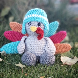 Puede incluir: Un pavo de ganchillo gris que lleva un casco azul y blanco con una papada roja. El pavo tiene una cola rosa, naranja y azul.