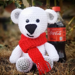 May include: White crocheted polar bear wearing a red scarf with a fringe. The bear is sitting on a bed of brown leaves and next to a bottle of Coca-Cola.