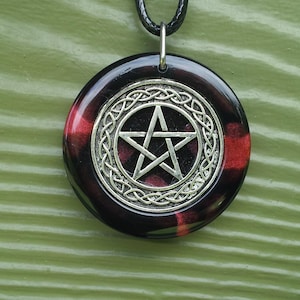 May include: A round pendant with a silver pentagram design, encircled by a silver Celtic knot pattern. The pendant has a black and red marbled background and hangs from a black cord.