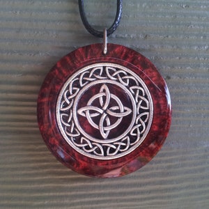 May include: A round pendant necklace with a silver Celtic knot design on a red background. The pendant is made of resin and has a black cord.