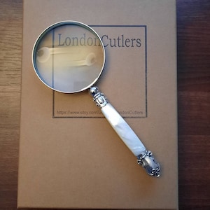 以下が含まれることがあります： 白い真珠貝のハンドルが付いた銀製の虫眼鏡。虫眼鏡は、"London Cutlers"というテキストとウェブサイトアドレスが書かれた茶色の箱の上に置かれています。