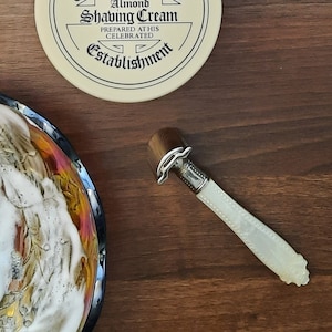 May include: A shaving brush with white lather in a decorative bowl, a safety razor with a white pearl handle, and a round container of shaving cream with the text 'Almond Shaving Cream Prepared at this Celebrated Establishment'.