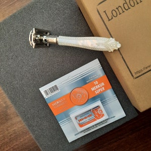 May include: A silver safety razor with a white pearlescent handle. The razor is next to a package of 10 Merkur Super Platinum Rostfrei-Stainless razor blades. The package is orange and white and has the text '10 Merkur Super' on it. The razor and blades are on a gray surface.
